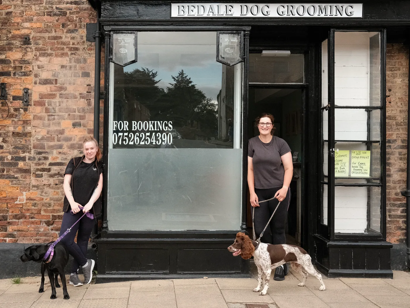 The black shopfront of Bedale Dog Grooming on Market Place, with Emma and Liv outside holding a springer spaniel and a black labrador